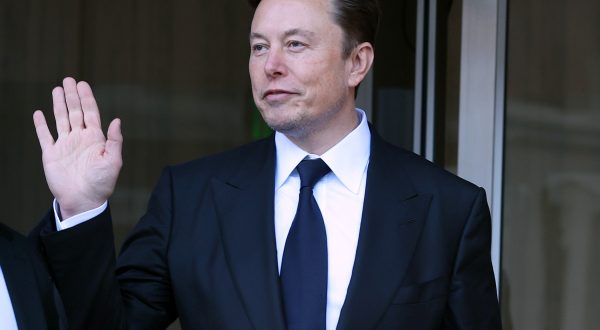Elon Musk in schwarzem Anzug grüßt mit seiner Hand