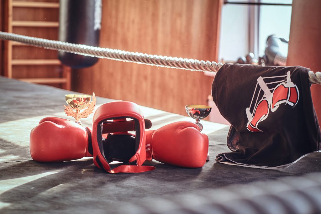 Rote Boxhandschuhe und ein Gesichtsschutz liegen vor zwei goldenen Pokalen