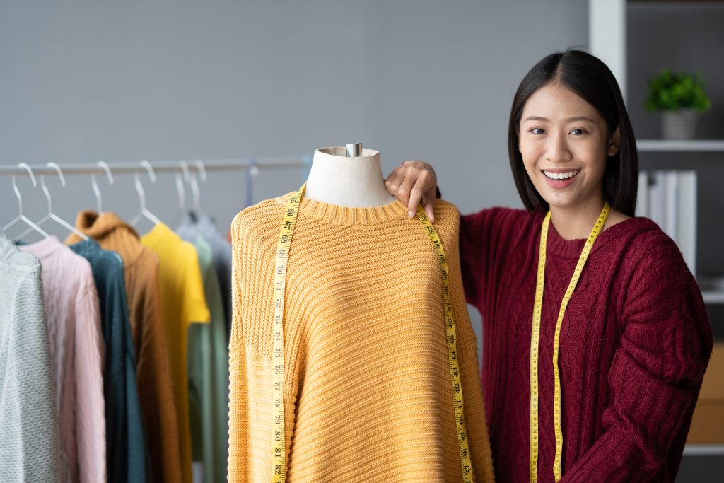 Asiatische Frau mit Maßband um den Hals steht vor Kleiderstange und neben Kleiderpuppe