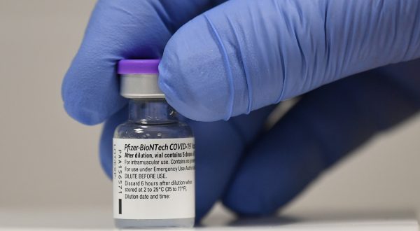 Hand mit blauem Handschuh greift auf ein Fläschchen mit Corona-Impfstoff von BionTech