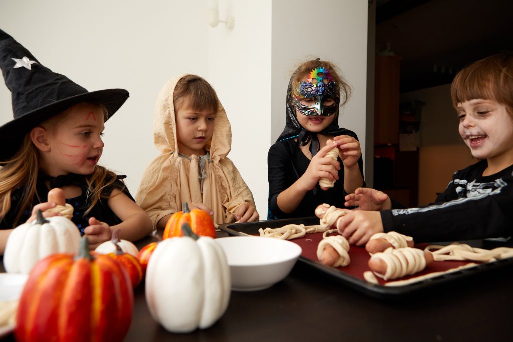 Vier Kinder sitzen verkleidet mit Hüten, Mantel und Maske an einem Tisch und wickeln Würstchen mit einem Teig ein. Vor ihnen viele kleine Kürbisse