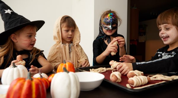 Vier Kinder sitzen mit einem Hut, Mantel, Schminke an einem Tisch und wickeln Teig um Würstchen. Vor ihnen liegen viele Kürbisse auf dem Tisch