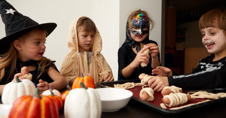 Vier Kinder sitzen mit einem Hut, Mantel, Schminke an einem Tisch und wickeln Teig um Würstchen. Vor ihnen liegen viele Kürbisse auf dem Tisch