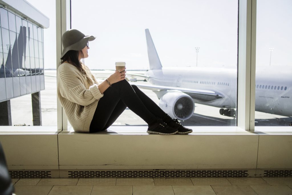 Frau mit Hut schaut zu einem Flugzeug und hält einen Papierbecher in der Hand.