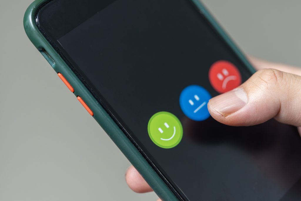 Handy in Nahaufnahme das drei Smiley in grün blau und rot zeigt. Der grüne lächelt freundlich, der blaue ist neutral und der rote ist negativ. Ein Daumen drückt auf eines der Symbole