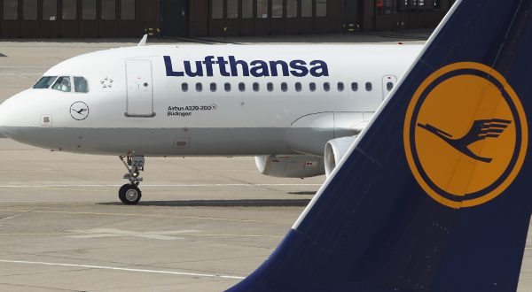 Flugzeugt der Lufthansa im Hintergrund. Davor der Flügel einer Lufthansa-Maschine in blau mit dem gelben Logo indem ein Kranich zu sehen ist