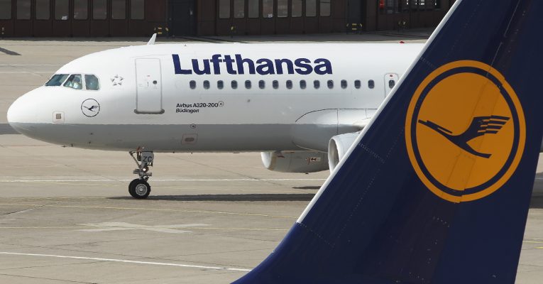 Flugzeugt der Lufthansa im Hintergrund. Davor der Flügel einer Lufthansa-Maschine in blau mit dem gelben Logo indem ein Kranich zu sehen ist