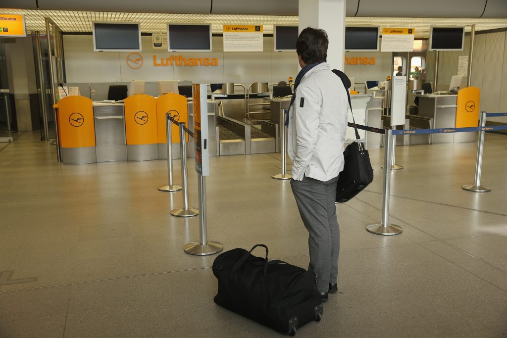 Fluggast mit einer umgehängten Notebook-Tasche und einer schwarzen Reisetasche steht vor dem leeren Check-In-Schalter der Lufthansa