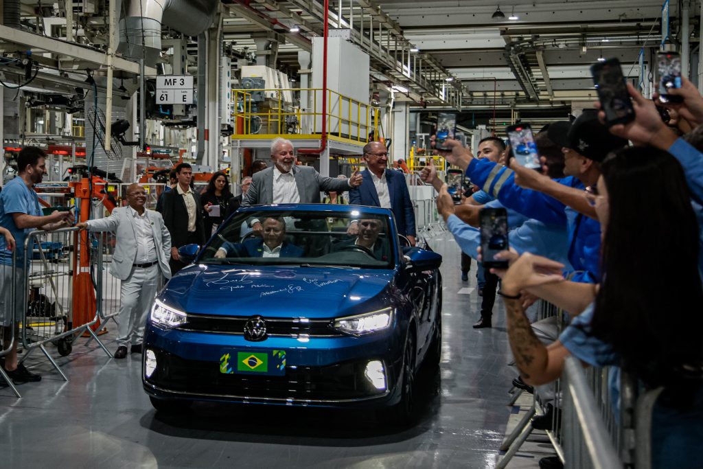 VW in blau in einer Werkshalle. Im Auto sind vier Menschen bei geöffnetem Dach zu sehen. Daneben viele Mitarbeiter die mit ihrem Handy fotografieren