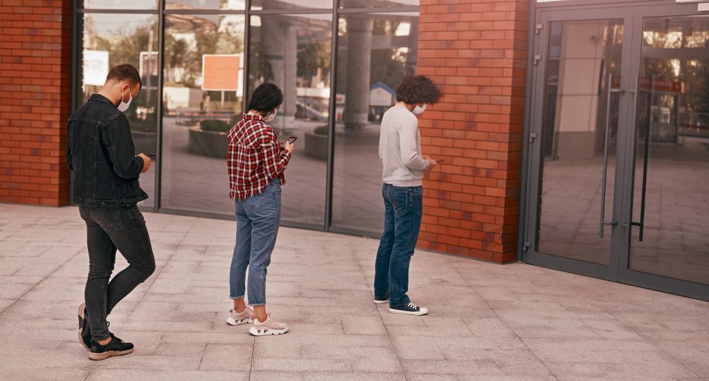 Drei Menschen stehen mit dem Handy in der Hand und Mundschutzmasken vor dem Eingang zu einem Shop