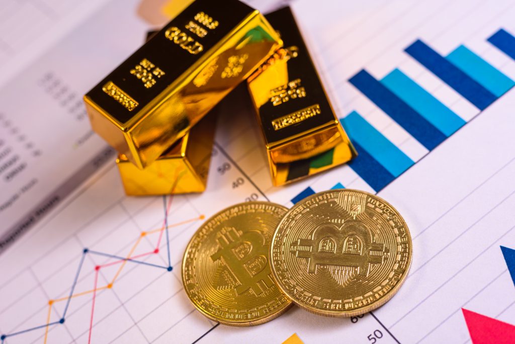 Drei Goldbarren und zwei Goldmünzen liegen auf einem Tisch auf einem Blatt Papier dass die Entwicklung des Goldpreises zeigt