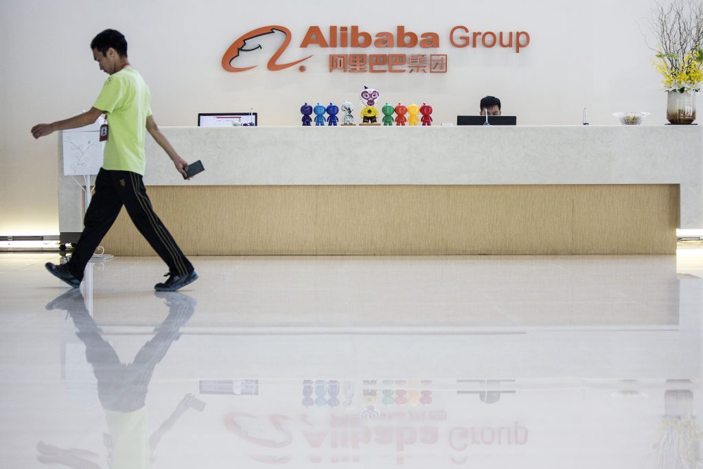 Logo der Alibaba Group an einem Empfang; davor viele unterschiedliche Plastikfiguren und ein Mann in hellgrünem T-Shirt und Jogginghose der nach links geht