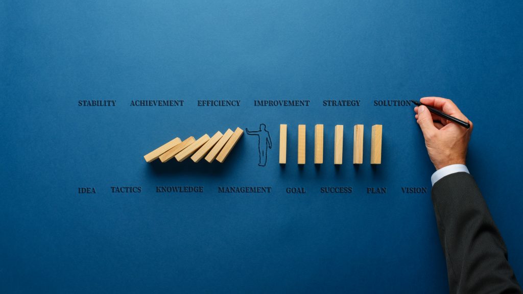 Blauer Hintergrund mit Worten wie Stability, Achievement, Efficiency, Improvement und weitere. In der Mitte ein  Umriss eines Menschen und sechs aufgestellte Holzklötze rechts sowie sechs Holzklötze im Fallen links und eine Hand mit einem Stift