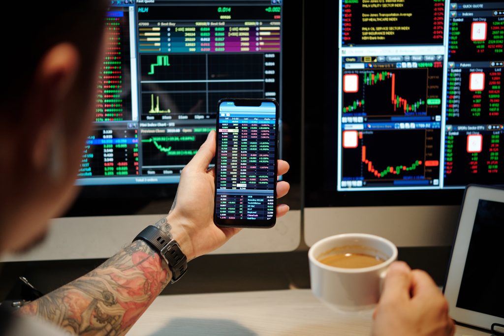 Mann hält ein Handy mit Aktienkursen und eine Tasse Kaffee vor zwei Monitore die auch Aktienkurse zeigen