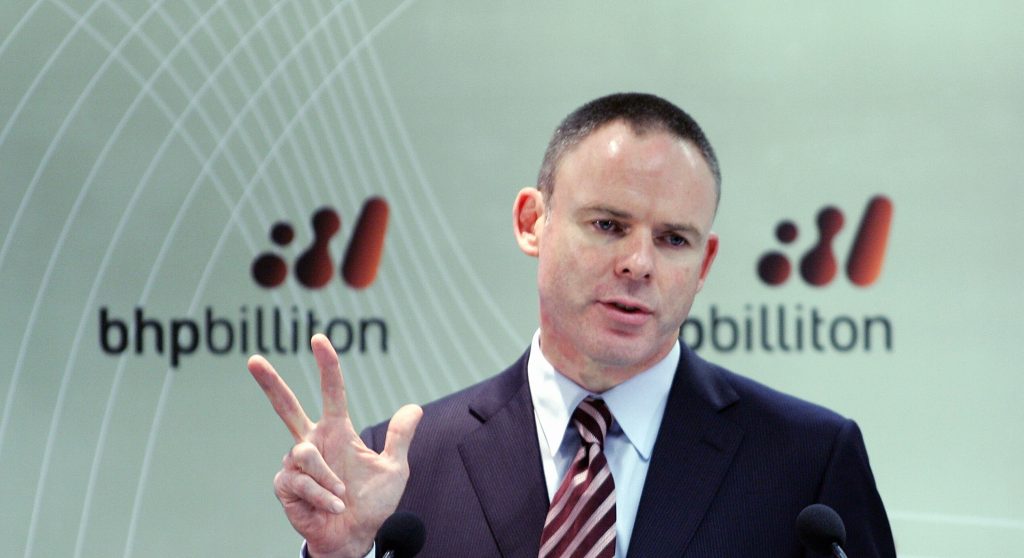 Mann mit Anzug und Krawatte hält drei Finger gespreizt hoch. Hinter ihm das Logo von bhp billition