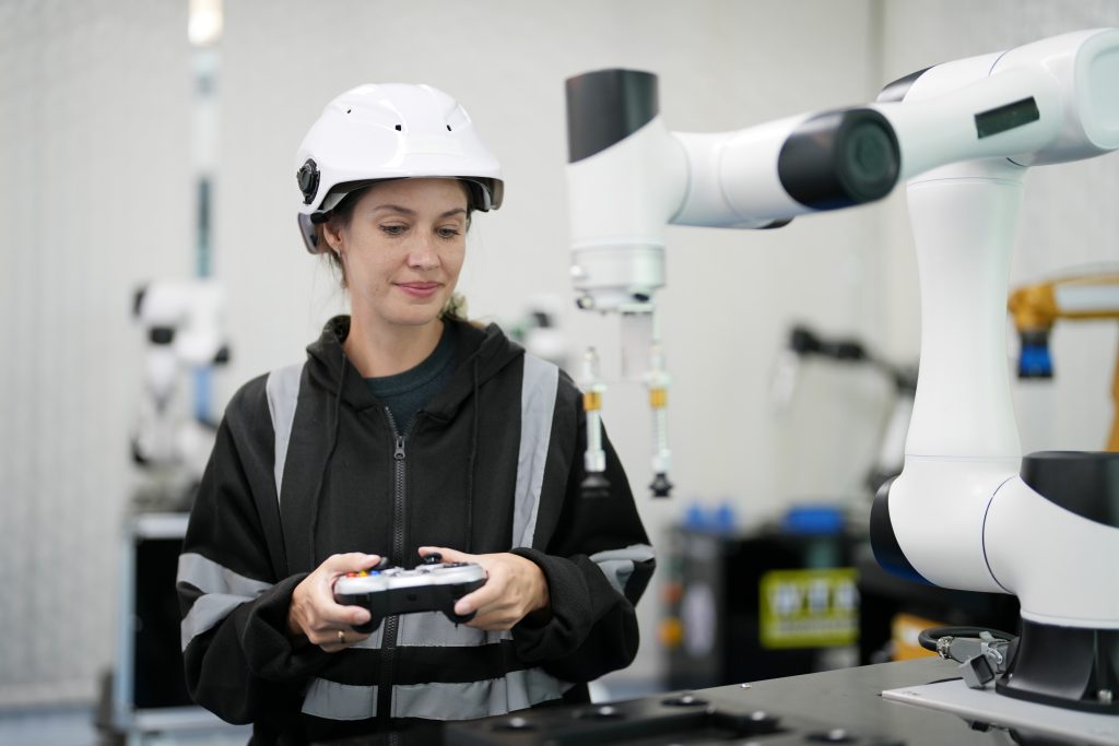 Frau steht mit einem weißen Schutzhelm und einem elektronischem Steuerungsgerät vor einem weißen Roboterarm