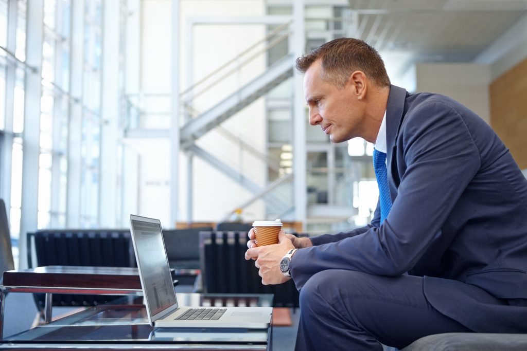 Mann mit blauem Anzug hält einen Becher Kaffee und blickt auf einen PC vor dem er sitzt